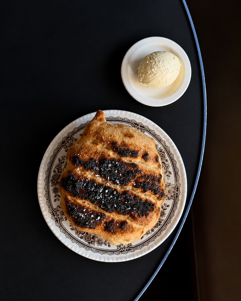 A round loaf of Sonny's bread, blistered and charred in stripes from the grill and scattered with sea salt, on a floral plate beside a scoop of whipped butter.